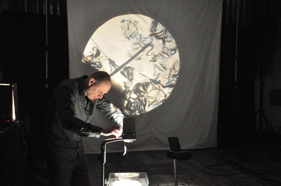 Mężczyzna pochylony nad rzutnikiem tworzy cienie i obrazy, które wyświetlają się na białym ekranie. Światło podkreśla jego skupienie i pracę twórczą.