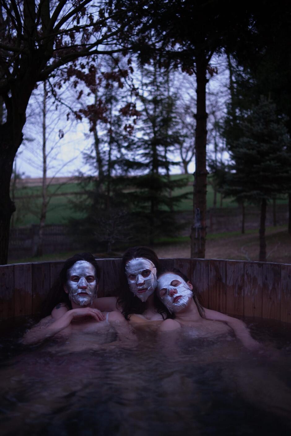 Trzy kobiety z maseczkami na twarzach siedzą w drewnianej balii z wodą, otoczone zimowym ogrodem. Zdjęcie jest tajemnicze i nieco surrealistyczne.