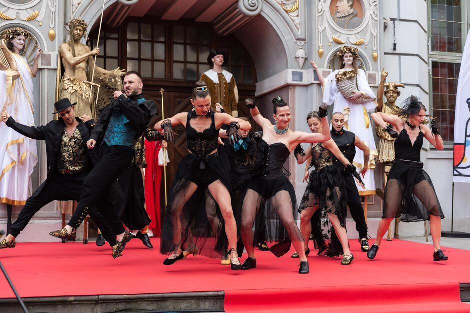 Na zdjęciu grupa tancerzy w efektownych, czarno-złotych kostiumach wykonuje dynamiczny układ choreograficzny na czerwonym dywanie przed zabytkowym budynkiem. Ich energiczne ruchy i ekspresja tworzą pełne pasji widowisko, wpisujące się w festiwalowy klimat wydarzenia.
