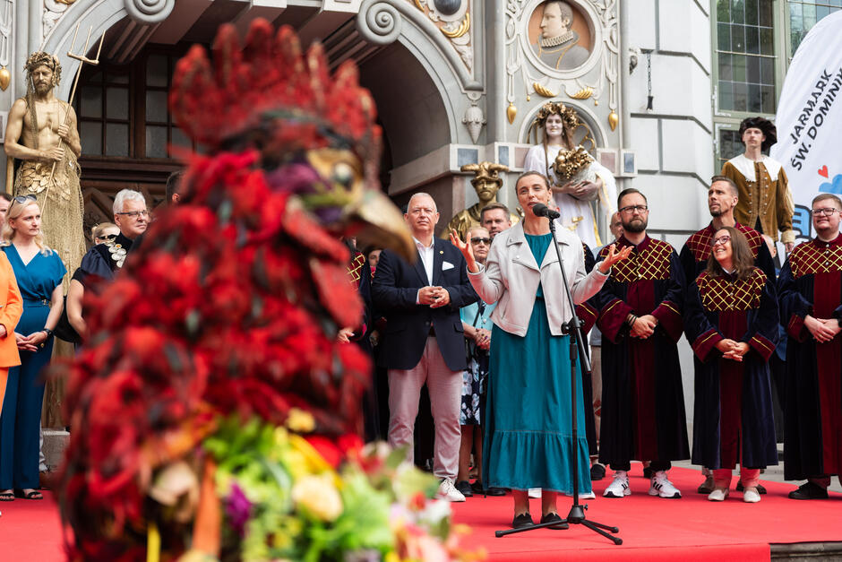 Na zdjęciu kobieta przemawia do mikrofonu podczas oficjalnego otwarcia wydarzenia, otoczona przez przedstawicieli władz i osoby w historycznych strojach. Na pierwszym planie widoczna jest barwna, symboliczna postać koguta z kwiatami, co dodaje uroczystości barwnego i tradycyjnego charakteru.
