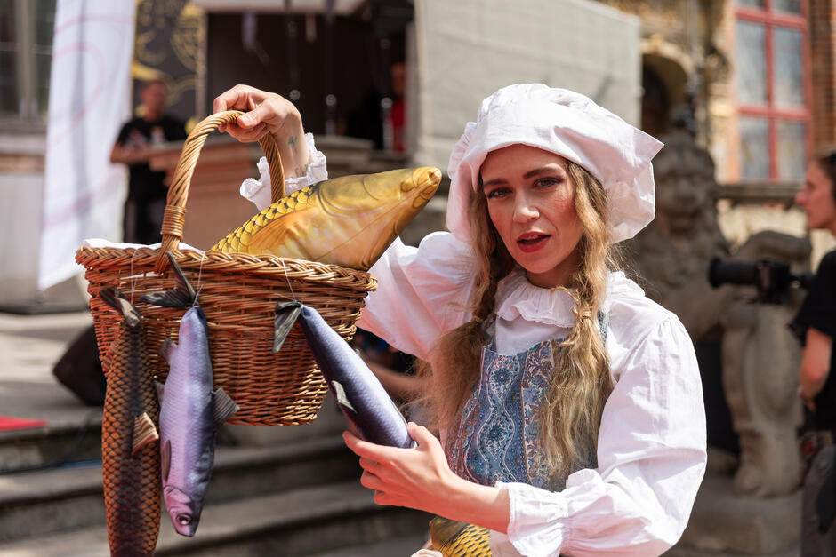 Na zdjęciu kobieta w stylizowanym stroju ludowym prezentuje wiklinowy kosz wypełniony sztucznymi rybami, wcielając się w postać tradycyjnej rybaczki. Jej ekspresyjny wyraz twarzy i kolorowy kostium dodają humorystycznego i teatralnego charakteru całej scenie.
