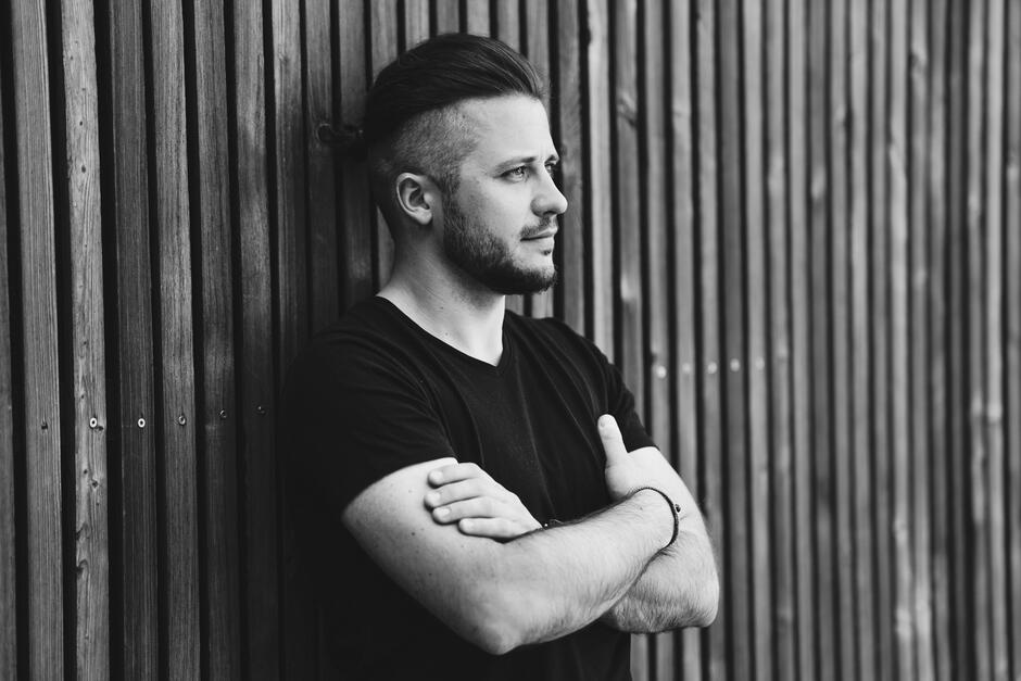 Na zdjęciu mężczyzna w czarnej koszulce, oparty o drewnianą ścianę z desek.