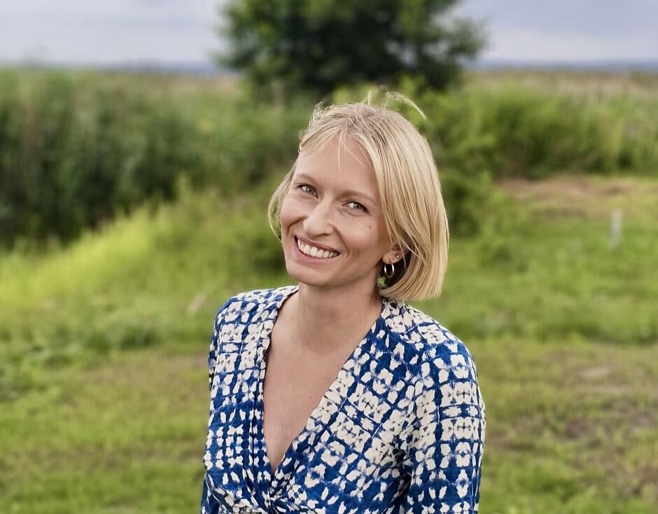 Na zdjęciu uśmiechnięta kobieta o blond włosach, na tle zieleni.