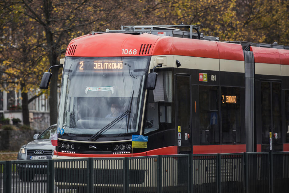 Na zdjęciu widoczny jest czerwono-czarny tramwaj linii 2 w Gdańsku, jadący w kierunku Jelitkowa. Z przodu pojazdu umieszczona jest mała flaga Ukrainy oraz szalik z napisem „Lechia Gdańsk”.