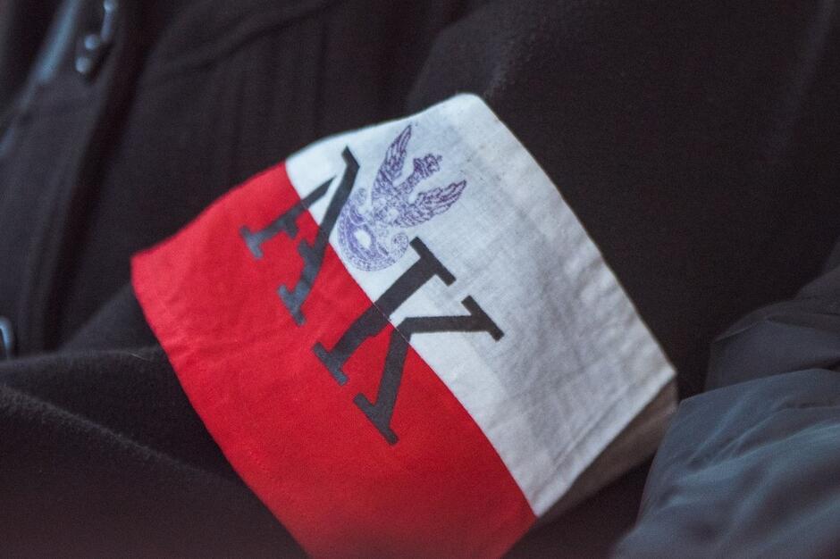 Biało-czerwona opaska, na niej odbite z pieczęci dwie duże czarne litery AK i powyżej mały orzełek w koronie