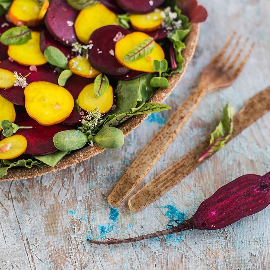 Na zdjęciu widoczna jest kolorowa sałatka z plasterkami czerwonych i żółtych buraków, ułożona na liściach sałaty i udekorowana świeżymi ziołami. Obok leży drewniany widelec i nóż oraz surowy burak, co podkreśla naturalny i zdrowy charakter dania. 