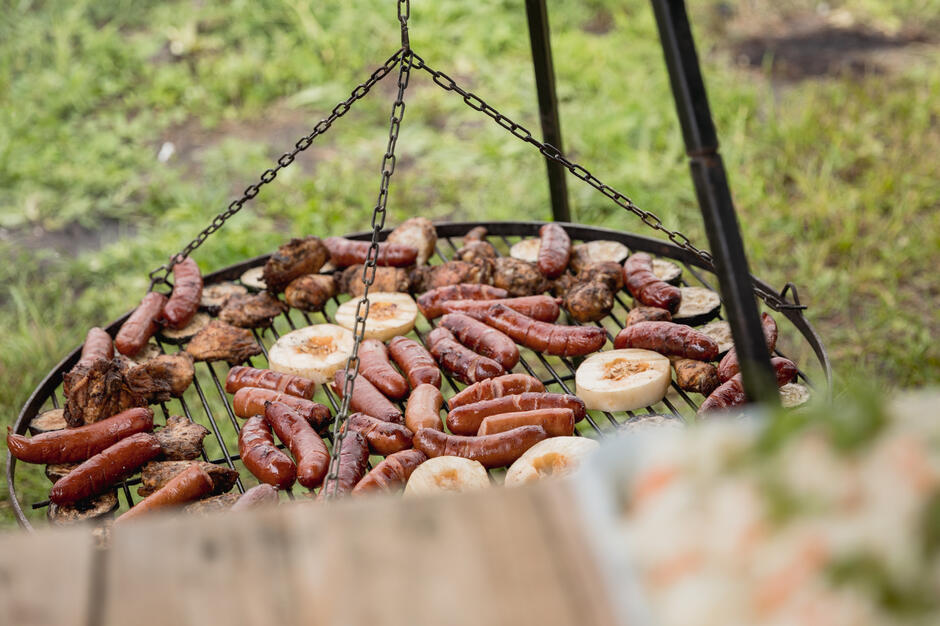 Na zdjęciu widać tradycyjnego grilla ogrodowego z rusztem zawieszonym na łańcuchach, na którym smażą się kiełbasy, kawałki mięsa i plastry warzyw, prawdopodobnie bakłażana lub cukinii. Całość odbywa się na świeżym powietrzu, co nadaje scenie piknikowego i biesiadnego klimatu. 