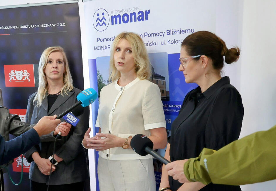 Trzy kobiety stoją podczas konferencji prasowej, przed nimi ręce trzymające mikrofony.