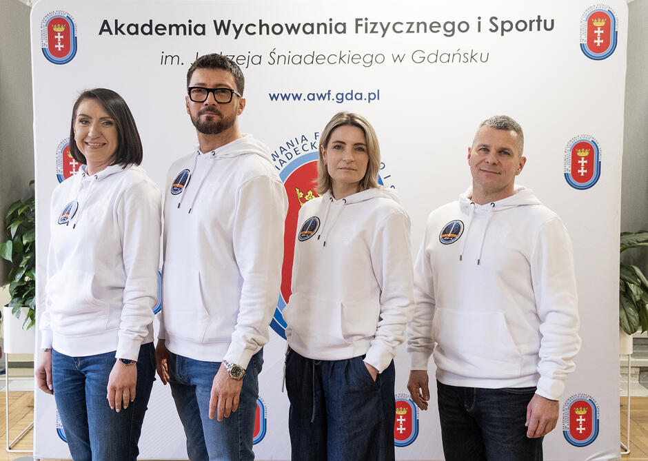 Cztery osoby pozują do zdjęcia na tle ścianki Akademii Wychowania Fizycznego i Sportu w Gdańsku - dwie kobiety i dwóch mężczyzn.