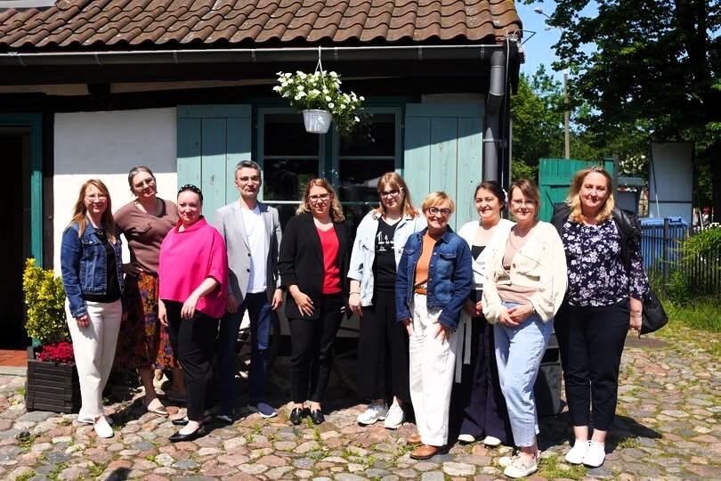 Na zdjęciu znajduje się grupa uśmiechniętych osób stojących przed urokliwym budynkiem z niebieskimi okiennicami i kwiatami, co sugeruje przyjazną atmosferę spotkania. Fotografia została wykonana podczas lipcowego spotkania realizatorów projektu „DAJ SPOKÓJ – środowiskowe wsparcie w pieczy zastępczej”, organizowanego przez Gdańską Fundację Innowacji Społecznej.
