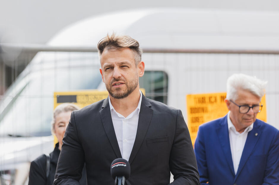 Na zdjęciu mężczyzna w garniturze przemawia do mikrofonu podczas wydarzenia plenerowego, prawdopodobnie konferencji prasowej, na tle ogrodzenia budowy i tablic informacyjnych. W tle widać inne osoby, co sugeruje udział przedstawicieli władz lub organizatorów związanych z inwestycją lub remontem.