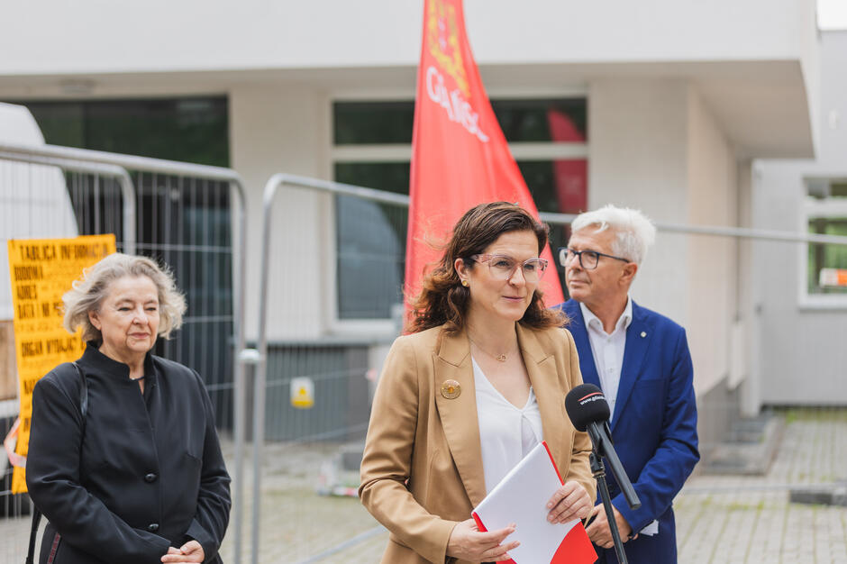 Na zdjęciu kobieta w beżowej marynarce przemawia do mikrofonu podczas konferencji prasowej, trzymając w rękach dokumenty; za nią widoczna jest czerwona flaga z napisem „Gdańsk”. W tle stoją dwie osoby, a całość odbywa się na tle ogrodzenia placu budowy, co sugeruje temat związany z miejską inwestycją lub remontem.
