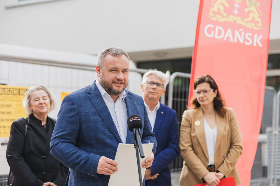 Na zdjęciu mężczyzna w niebieskiej marynarce przemawia podczas konferencji prasowej, trzymając dokumenty i stojąc przed mikrofonem z logo „gdansk.pl”. W tle znajdują się trzy inne osoby, a całość odbywa się na tle budynku i czerwonej flagi z herbem oraz napisem „GDAŃSK”, co sugeruje oficjalne wydarzenie miejskie, prawdopodobnie dotyczące inwestycji lub remontów.
