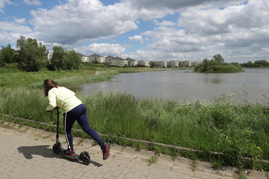 na zdjęciu dziewczynka na hulajnodze, za nią duży staw wypełniony wodą