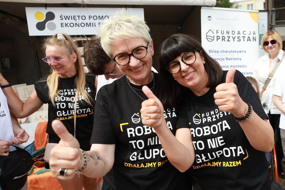 Na zdjęciu uśmiechnięte kobiety w koszulkach z hasłem „Robota to nie głupota – działajmy razem” pozują z uniesionymi kciukami podczas wydarzenia „Święto Pomorskiej Ekonomii Społecznej”. W tle widoczne są stoiska i banery Fundacji Przystań, promujące działania społeczne i charytatywne.