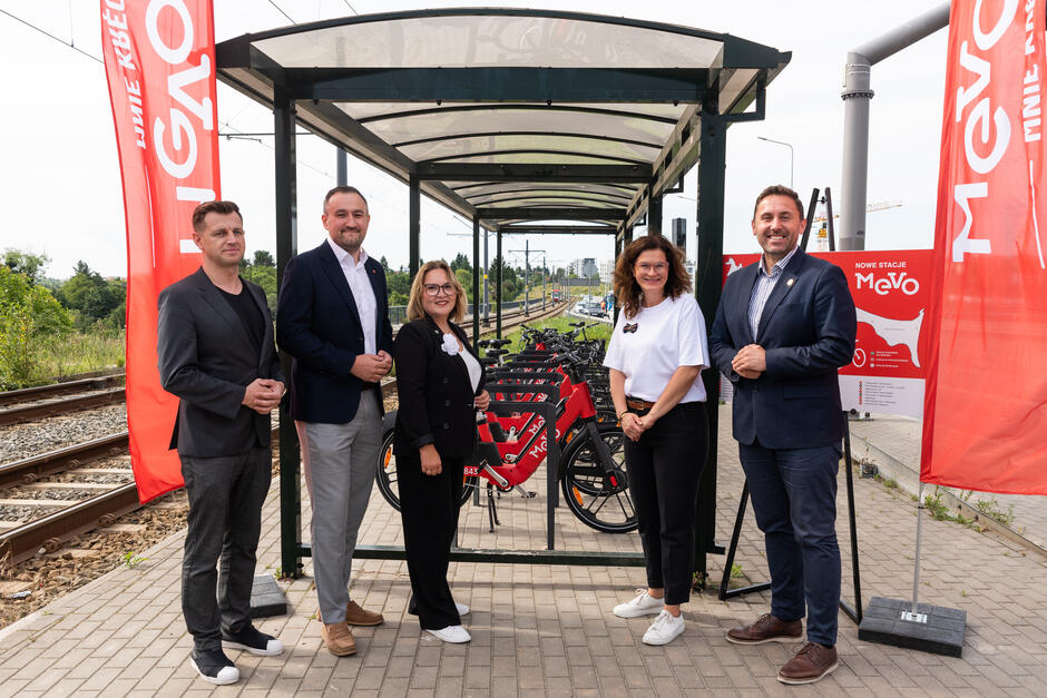 Na zdjęciu znajduje się grupa sześciu osób pozujących przy nowo otwartej stacji rowerów miejskich MEVO, zlokalizowanej przy torach kolejowych. W tle widoczne są zaparkowane czerwone rowery elektryczne oraz banery i tablica informacyjna promujące system MEVO.
