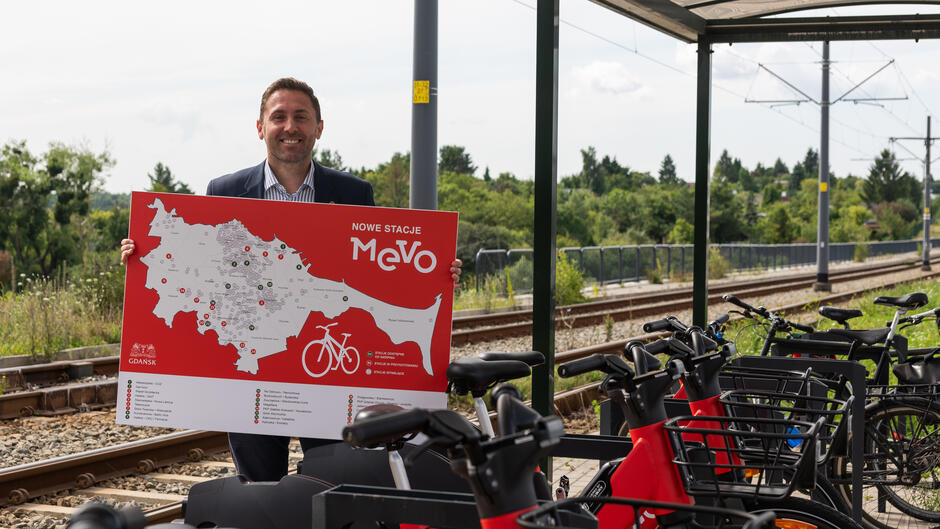 Na zdjęciu mężczyzna w garniturze trzyma dużą planszę z mapą nowych stacji rowerowych MEVO, stojąc przy torach kolejowych i zadaszonej stacji rowerowej. W tle widoczne są zaparkowane czerwone rowery elektryczne oraz zielone otoczenie, co sugeruje inaugurację nowej infrastruktury rowerowej.
