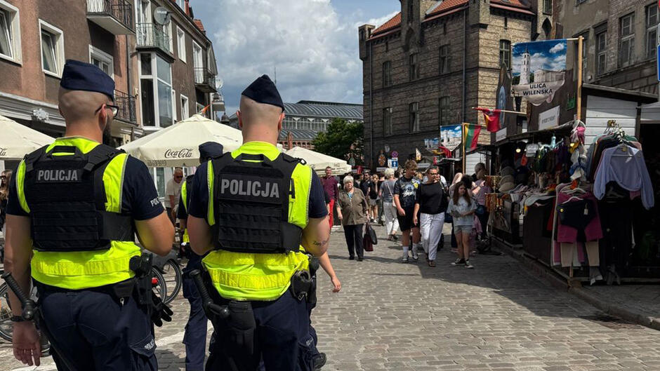 Na zdjęciach widać kilku policjantów patrolujących ruchliwą, brukowaną uliczkę miejską podczas imprezy plenerowej lub jarmarku. W tle tłum ludzi spaceruje między stoiskami z odzieżą i pamiątkami.