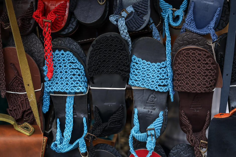 Na zdjęciu widoczne są ręcznie wykonane sandały z plecionymi, kolorowymi paskami — dominują barwy niebieska, czarna, czerwona i brązowa. Obuwie ma solidne, skórzane podeszwy i prezentowane jest na targowym stoisku w estetycznym, uporządkowanym układzie. 