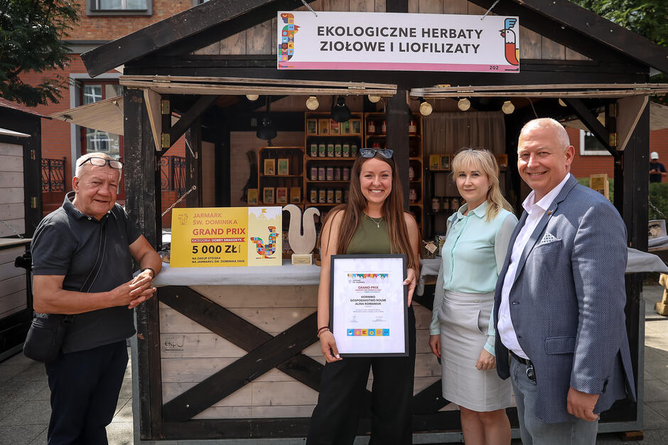 Na zdjęciu grupa osób pozuje przed stoiskiem z ekologicznymi herbatami ziołowymi i liofilizatami, które zdobyło nagrodę Grand Prix w kategorii Dobry Smakołyk  na Jarmarku św. Dominika. Uśmiechnięta kobieta trzyma dyplom, a na ladzie widoczny jest symboliczny czek na 5000 zł oraz kolorowa statuetka.
