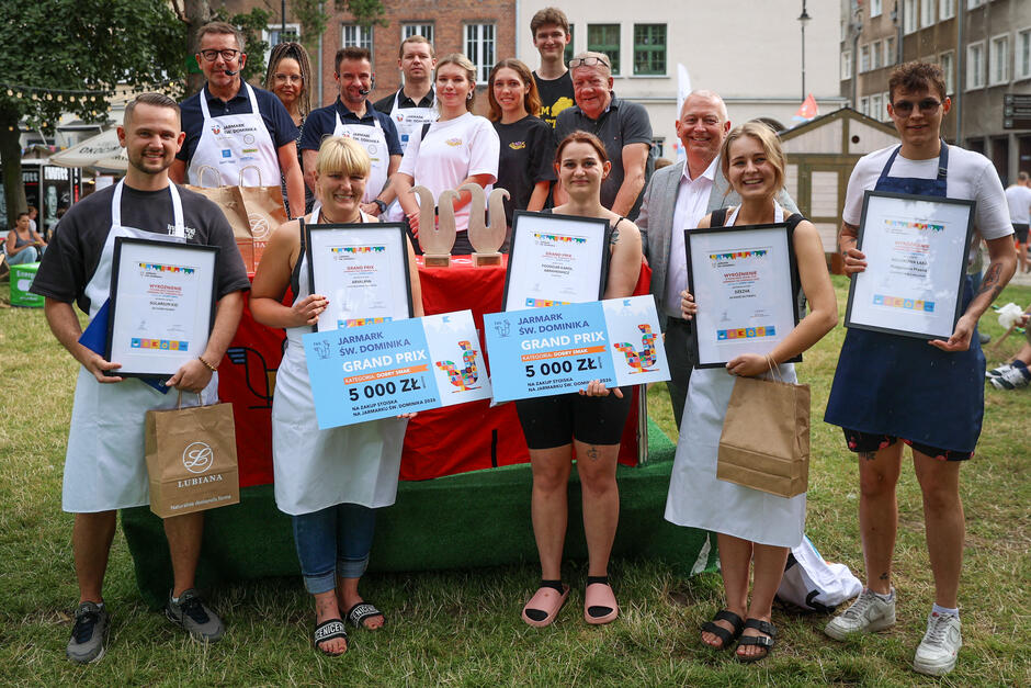 Na zdjęciu widać grupę laureatów konkursu kulinarnego z Jarmarku św. Dominika, dumnie prezentujących dyplomy i symboliczne czeki o wartości 5000 zł w kategorii „Dobry Smak”. Uczestnicy pozują z uśmiechem na tle zielonej trawy i miejskiej scenerii, a w centralnym punkcie widoczne są dwie statuetki w kształcie jarmarcznych symboli.
