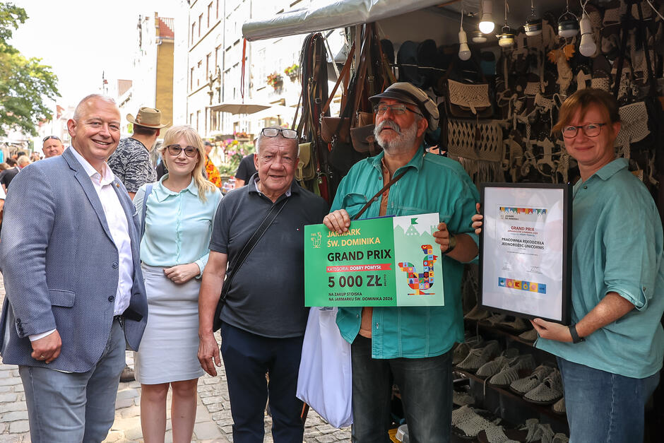 Na zdjęciu grupa uśmiechniętych osób pozuje przed stoiskiem rzemieślniczym na Jarmarku św. Dominika, trzymając nagrody – czek na 5000 zł oraz dyplom za zdobycie Grand Prix w kategorii Dobry Pomysł . W tle widoczne są ręcznie wykonane wyroby, takie jak torebki i sandały, co podkreśla charakter nagrodzonego rzemiosła. 