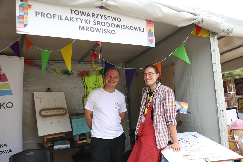 Na zdjęciu widzimy stoisko Towarzystwa Profilaktyki Środowiskowej Mrowisko , ozdobione kolorowymi proporczykami, z uśmiechniętym mężczyzną i kobietą stojącymi przed wejściem. Atmosfera jest przyjazna i otwarta, a elementy takie jak tęczowa flaga i plansze edukacyjne sugerują działania na rzecz społeczności i integracji. 