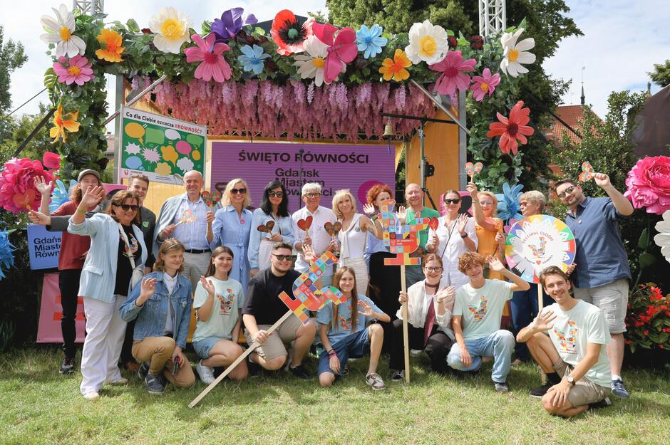 Na zdjęciu grupa osób pozuje radośnie przed sceną udekorowaną kwiatami podczas wydarzenia Święto Równości  w Gdańsku. Uczestnicy trzymają kolorowe symbole równości i serca, wyrażając wsparcie dla idei otwartości, różnorodności i integracji społecznej. 