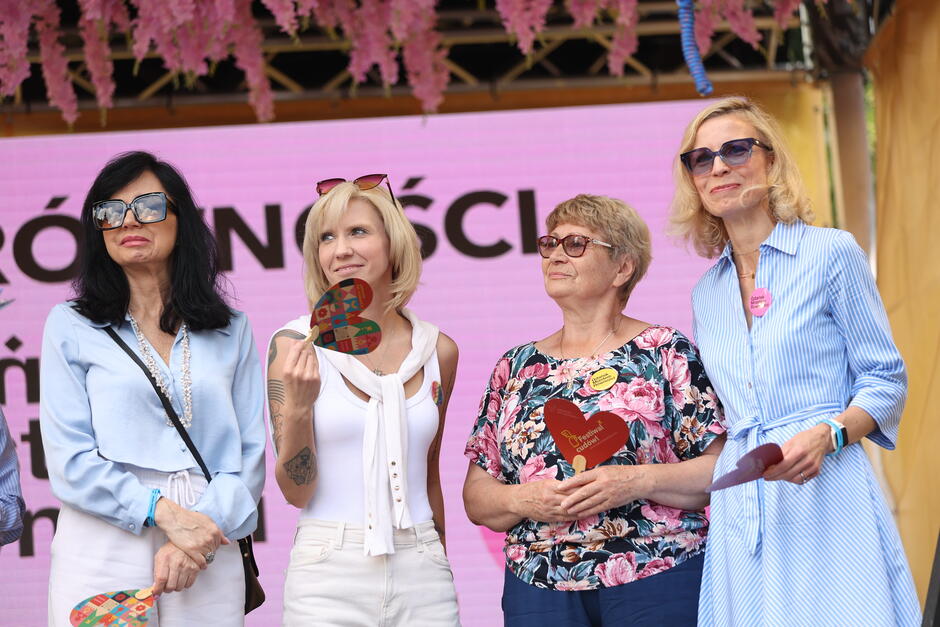 Na zdjęciu cztery kobiety stoją na scenie podczas wydarzenia „Święto Równości” w Gdańsku, trzymając kolorowe serca symbolizujące różnorodność i solidarność. Ich uśmiechy i zaangażowanie podkreślają pozytywną atmosferę oraz ideę wspólnoty i akceptacji. 