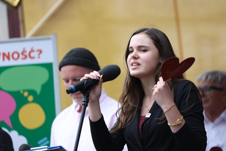 Na zdjęciu młoda kobieta przemawia do mikrofonu na scenie podczas wydarzenia, trzymając czerwone serce z napisem „Festival Równości Gdańsk”. Jej zaangażowanie i ekspresja świadczą o poruszającym i ważnym wystąpieniu, które towarzyszy celebracji wartości równości i otwartości. 