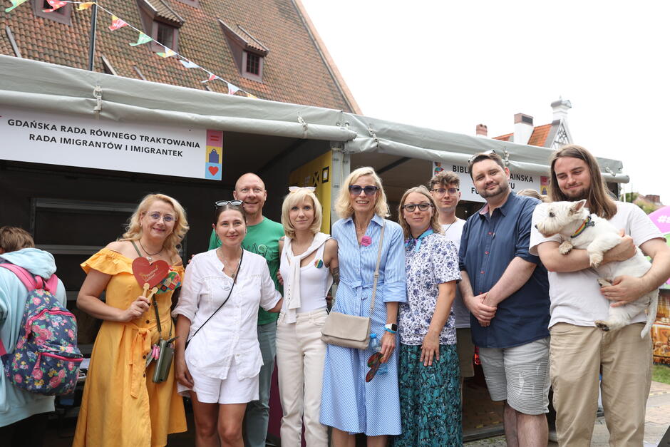 Na zdjęciu grupa osób pozuje wspólnie przed namiotem oznaczonym szyldem „Gdańska Rada Równego Traktowania – Rada Imigrantów i Imigrantek”. Uczestnicy wydarzenia uśmiechają się, trzymają kolorowe symbole równości, a wśród nich znajduje się również pies, co podkreśla przyjazną i otwartą atmosferę. 