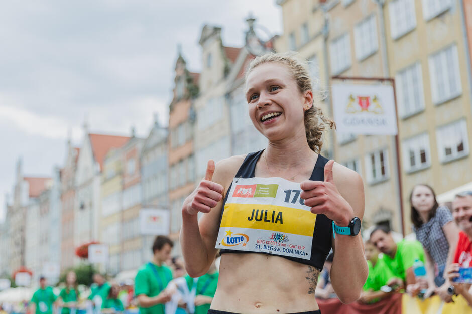 Uśmiechnięta biegaczka pokazuje kciuki w górę tuż po zakończeniu biegu ulicznego, wśród kibiców i na tle kolorowych kamienic. Na numerze startowym widać oznaczenia „Bieg Kobiet 5 km – GAT Cup” oraz „31. Bieg św. Dominika” w Gdańsku.

