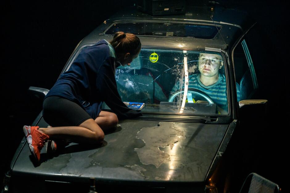 Dwoje aktorów w scenie z samochodem. Ona siedzi na masce auta. On jest w kokpicie, za kierownicą, przez szybę wpatruje się w nią