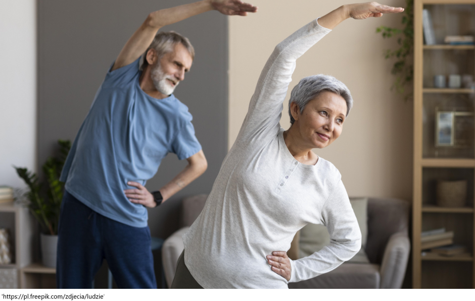 Starsza kobieta i starszy mężczyzna wykonują wspólnie ćwiczenia rozciągające w domowym zaciszu. Kobieta na pierwszym planie ma krótkie siwe włosy i jasnoszarą bluzkę, unosi jedno ramię nad głowę i pochyla tułów w bok, z uśmiechem na twarzy. Mężczyzna w tle ubrany jest w niebieski T-shirt i granatowe spodnie, wykonuje ten sam ruch. W tle widoczny jest przytulny salon z regałem, rośliną doniczkową i miękkimi fotelami. Scena ukazuje aktywność fizyczną seniorów w domowym środowisku.

Źródło zdjęcia: Freepik – zdjęcia ludzi