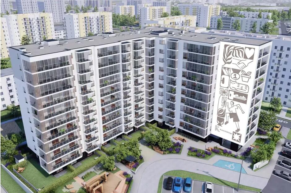 Na zdjęciu widoczny jest nowoczesny kompleks mieszkalny z dużymi balkonami i starannie zagospodarowanym dziedzińcem z placem zabaw oraz zielenią. Na bocznej elewacji budynku znajduje się mural przedstawiający symboliczne grafiki związane z codziennym życiem i społecznością.
