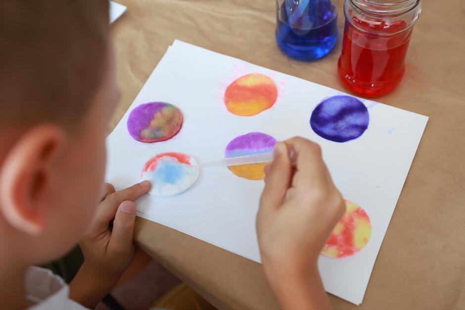 Dziecko wykonuje kolorowe prace plastyczne, nakładając pipetą barwniki na okrągłe płatki kosmetyczne ułożone na kartce. Obok stoją słoiczki z kolorowymi cieczami, co wskazuje na kreatywną zabawę połączoną z nauką mieszania kolorów. 