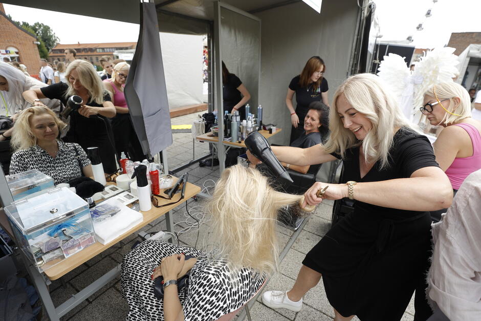 Stanowisko fryzjerskie na jarmarku w mieście. Fryzjerka czesze kobietę, a jej pracy przyglądają się inni ludzie.