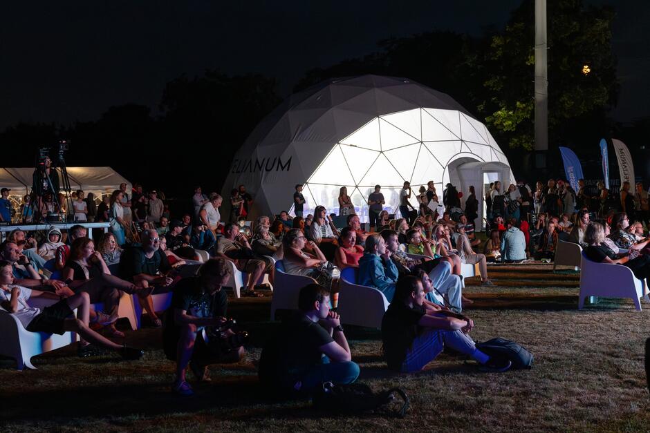 Na zdjęciu widać wieczorne wydarzenie plenerowe, gdzie liczna publiczność siedzi na leżakach i trawie, oglądając coś na scenie lub ekranie poza kadrem. W tle znajduje się podświetlony biały namiot kopułowy z napisem Hevelianum 