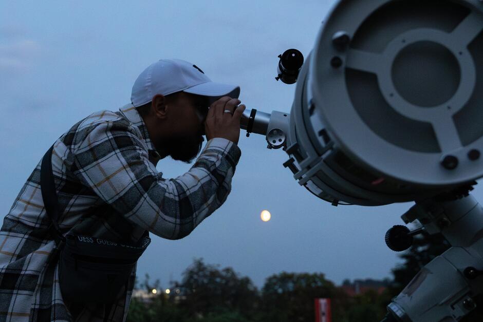 Zdjęcie przedstawia mężczyznę w czapce z daszkiem i koszuli w kratę, który obserwuje niebo przez duży teleskop. W tle widać wieczorne niebo z wyraźnie widocznym Księżycem unoszącym się nad linią drzew. 