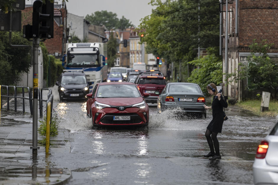 Na zdjęciu widać ulicę zalaną przez ulewę, po której przejeżdżają samochody, wzbijając wodę na boki. Z prawej strony młoda kobieta z parasolką w ręce próbuje ostrożnie przejść przez zalany chodnik. 