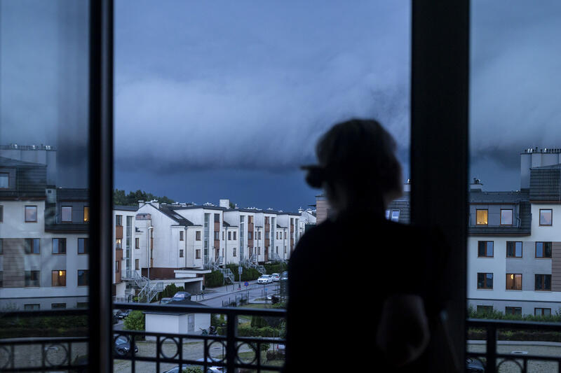 Na zdjęciu widzimy osobę obserwującą z balkonu nadciągającą burzę z charakterystycznym wałem szkwałowym na niebie. Ciemne chmury kontrastują z jasnymi fasadami bloków mieszkalnych, tworząc napiętą i groźną atmosferę. 
