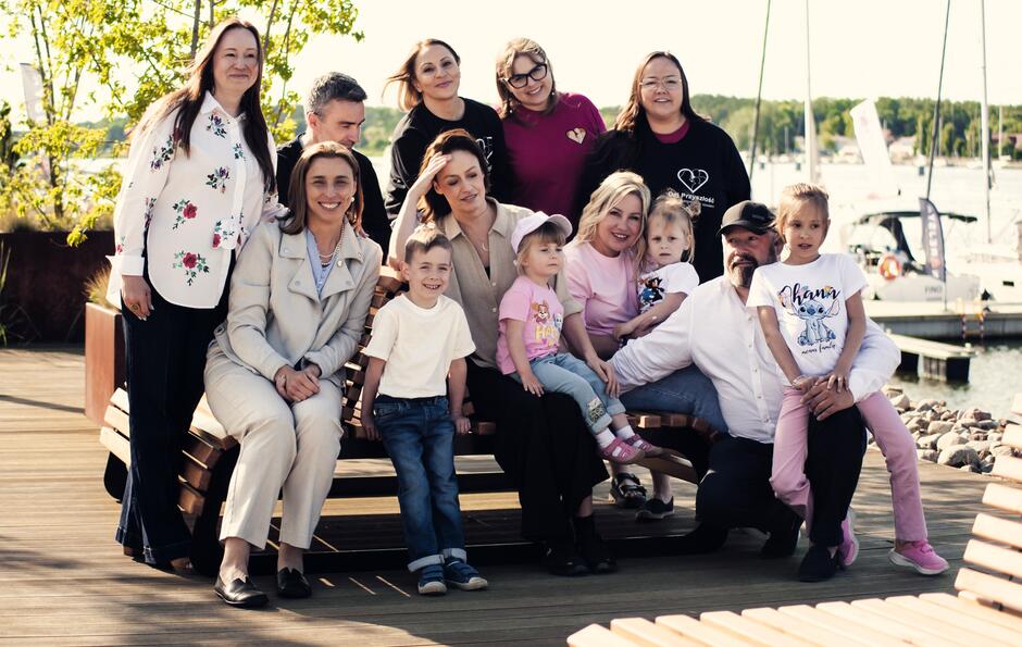 Kilka osób dorosłych i kilkoro dzieci pozuje do zdjęcia na ławce w przystani jachtowej.