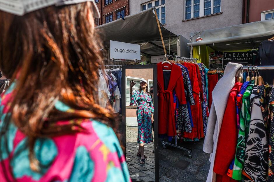 Na zdjęciu widzimy kobietę przymierzającą kolorowe ubranie na jarmarku, najprawdopodobniej podczas Jarmarku św. Dominika w Gdańsku. Stoi ona przed lustrem, w którym odbija się jej uśmiechnięta twarz oraz sylwetka w kwiecistej sukience. Nosi też czapkę z daszkiem. Za nią znajdują się stoiska z ubraniami, m.in. marki „angaretchka” oraz „Tabanna – Anna Tabaczyńska”. W tle widać kamienice i namioty wystawców. 