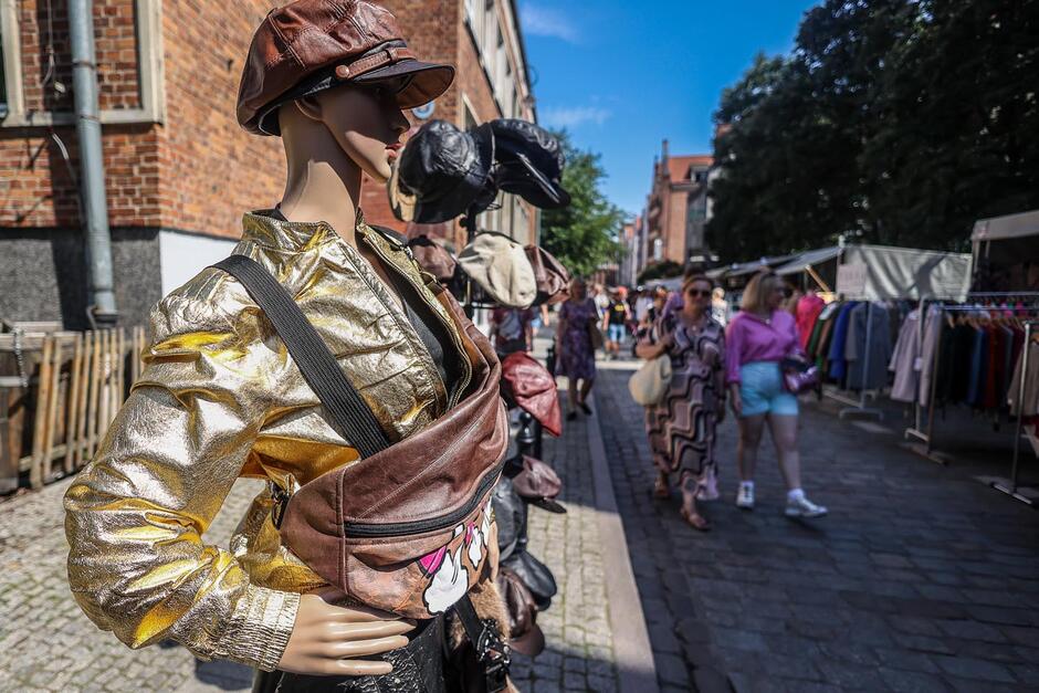 To kolejne zdjęcie z Jarmarku św. Dominika w Gdańsku, które oddaje klimat wydarzenia – pełnego koloru, mody i rękodzieła. Na pierwszym planie widzimy manekina ubranego w złotą, błyszczącą kurtkę, skórzaną czapkę i dużą nerkę (saszetkę) z nadrukiem. Obok niego widoczne są inne czapki wystawione na sprzedaż. W tle przechadzają się ludzie oglądający stoiska, w tym dwie kobiety – jedna w fioletowo-brązowej sukience w geometryczne wzory, druga w różowej koszuli i jasnych szortach. Po prawej stronie zdjęcia widać rzędy ubrań zawieszonych na wieszakach pod namiotami.