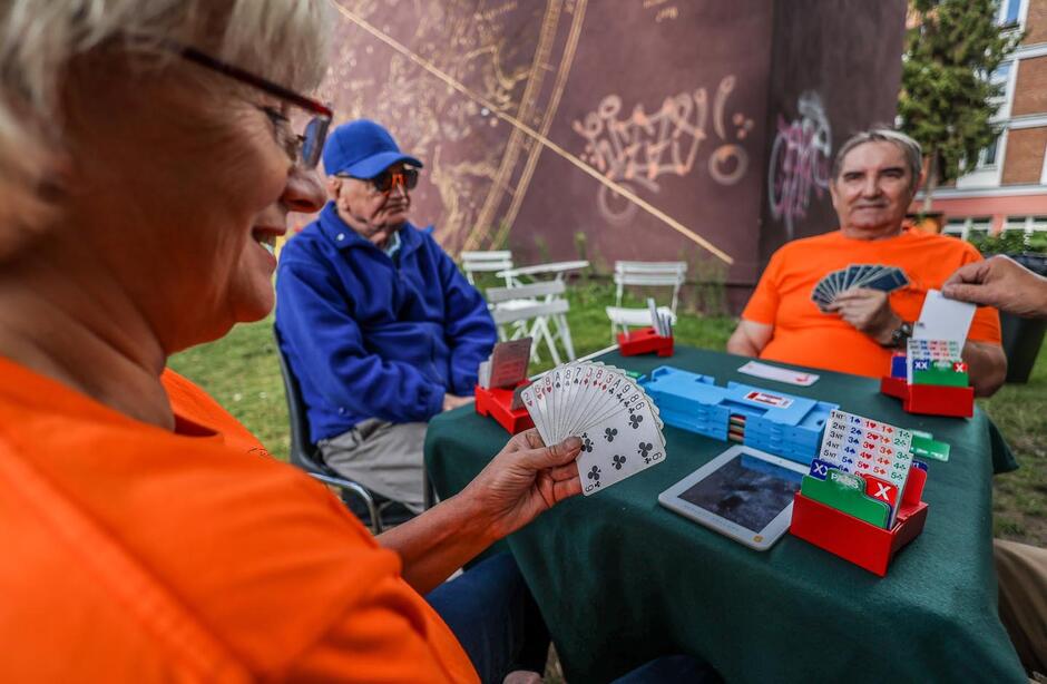 To zdjęcie przedstawia grupę starszych osób grających w brydża na świeżym powietrzu, prawdopodobnie podczas jednego z wydarzeń towarzyszących Jarmarkowi św. Dominika w Gdańsku.