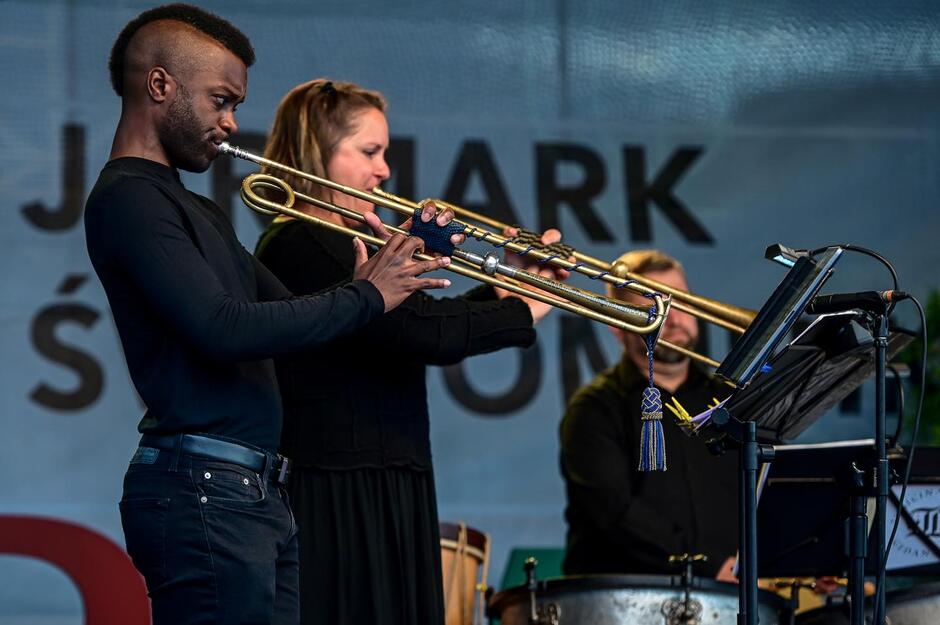 To zdjęcie przedstawia występ muzyczny na żywo podczas Jarmarku św. Dominika. Na pierwszym planie widzimy skupionego trębacza grającego z pełnym zaangażowaniem. Obok niego, nieco z tyłu, kobieta gra na puzonie. W tle, za nimi, siedzi perkusista z zestawem instrumentów i nutami. Cała trójka ubrana jest na czarno, co podkreśla powagę i profesjonalizm występu.