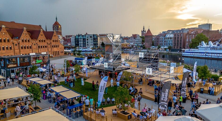 Na zdjęciu widać tętniące życiem nabrzeże Motławy w Gdańsku, gdzie odbywa się plenerowe wydarzenie z licznymi stoiskami, leżakami i odwiedzającymi. W tle można dostrzec charakterystyczne zabytkowe budynki Głównego Miasta oraz napisy „Port Gdańsk” i „Żegluga Gdańska”. 
