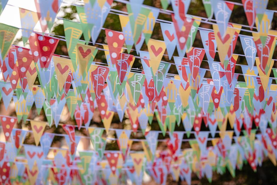 Na zdjęciu widoczne są kolorowe chorągiewki rozwieszone nad głowami uczestników wydarzenia plenerowego, ozdobione wzorami serc, ptaków, budynków i kropek. Barwna dekoracja tworzy radosną, festynową atmosferę i sugeruje obecność święta lub miejskiej imprezy. 