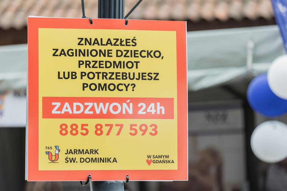 Na zdjęciu znajduje się tablica informacyjna z Jarmarku św. Dominika w Gdańsku, zawierająca numer telefonu alarmowego dostępnego 24 godziny na dobę. Służy ona do kontaktu w przypadku znalezienia zaginionego dziecka, przedmiotu lub potrzeby pomocy. 