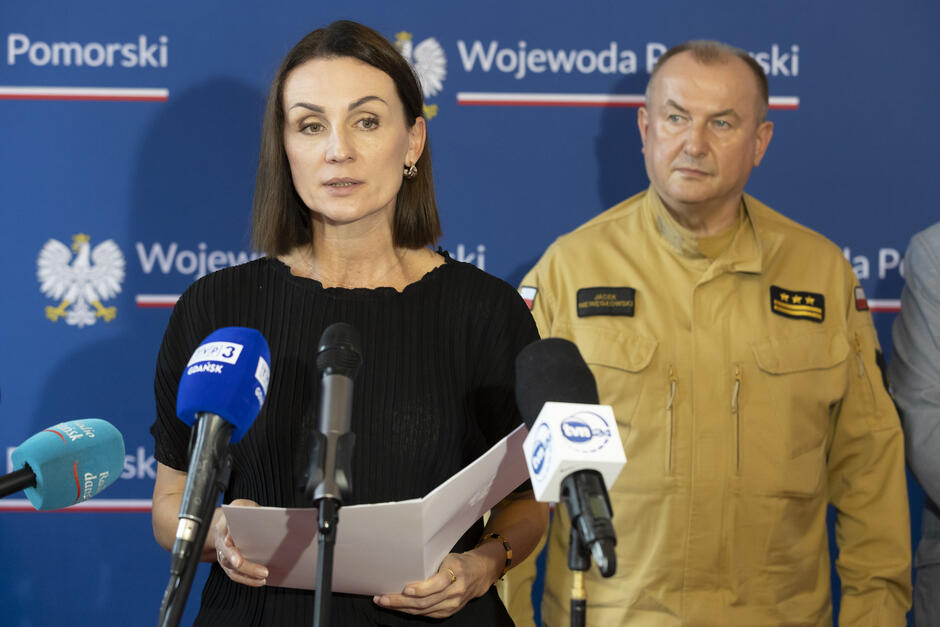 Kobieta i mężczyzna w mundurze strażaka stoją przed mikrofonem podczas konferencji prasowej. Kobieta mówi do mikrofonu.